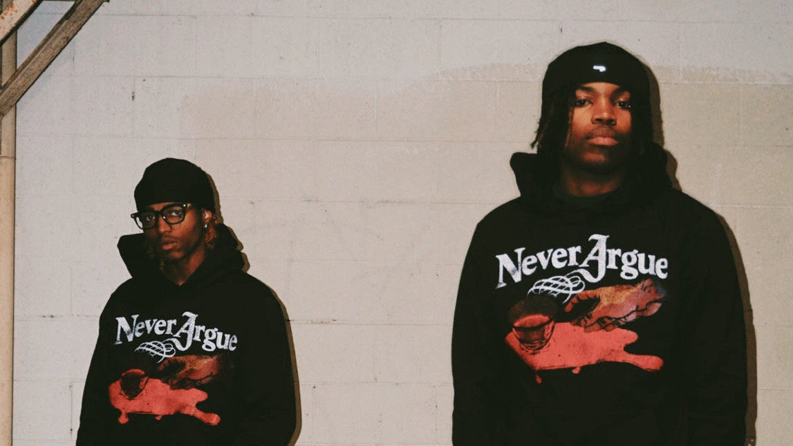 Two people wearing black hoodies with 'Never Argue' graphic design against a white brick wall.